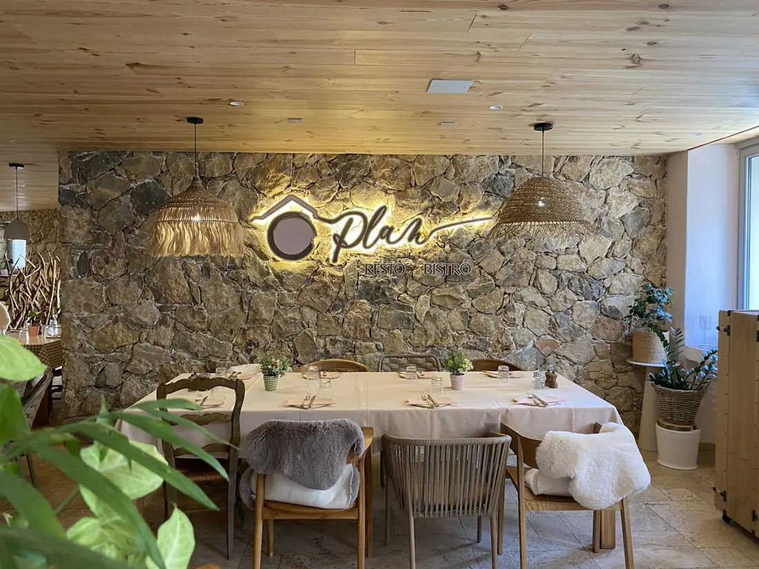 La salle d'Ô Plan : mur de pierre et enseigne lumineuse du logo, plafond en bois, grande table nappée